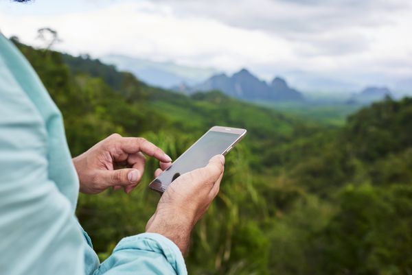 نزدیک یک دست یک مرد در حال نگه داشتن تلفن همراه با صفحه نمایش کپی در برابر تاری جنگل است مرد جوان در هنگام ماجراجویی تابستانی در تایلند در حال جستجوی اطلاعات از طریق تلفن همراه است