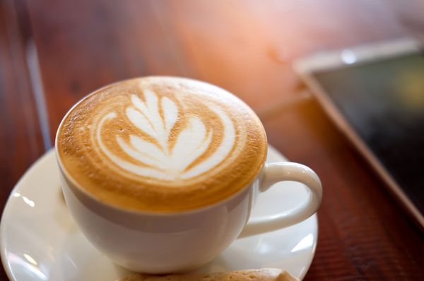 یک فنجان قهوه تازه در بشقاب سفید و گوشی هوشمند با زمینه چوبی با نور خورشید در صبح