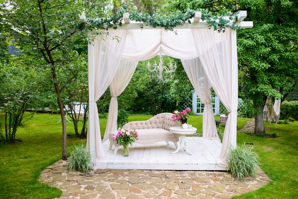 کاناپه ظریف تابستانی در باغ های سرسبز کاناپه کلاسیک مناسب با گلهای تزئین شده در گلدان با پرده های سفید در باغ تازه تزئین شده