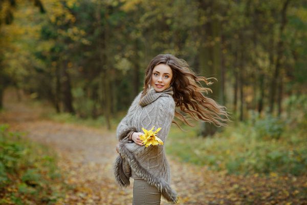 پرتره دختری بسیار زیبا و خندان در ژاکت گره دار موهایی که در پاییز در پارک پرواز می کنند