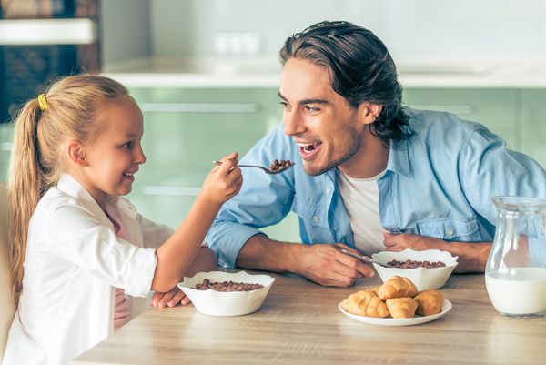 دختر بچه ناز و پدر خوش تیپش هنگام خوردن صبحانه در آشپزخانه در خانه لبخند می زنند دختر در حال تغذیه پدر خود با یک قاشق است