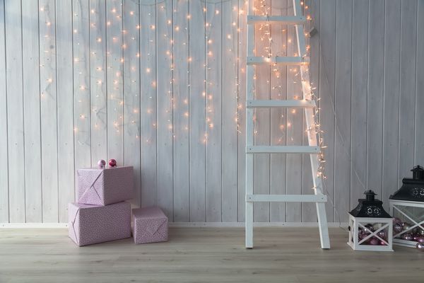 چراغ های کریسمس که روی یک زمینه چوبی سفید با جعبه های هدیه صورتی و پله ها می سوزند
