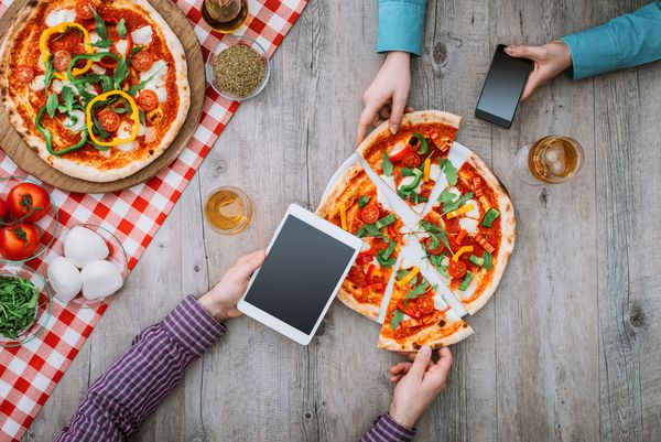 دوستانی که یک پیتزای خوشمزه در رستوران با هم دارند در حال خوردن و استفاده از یک تبلت و یک تلفن هوشمند هستند