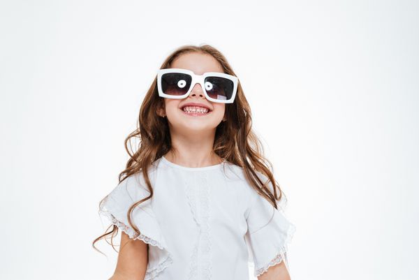 پرتره دختر بچه شاد در عینک آفتابی ایستاده و لبخند بر فراز زمینه سفید