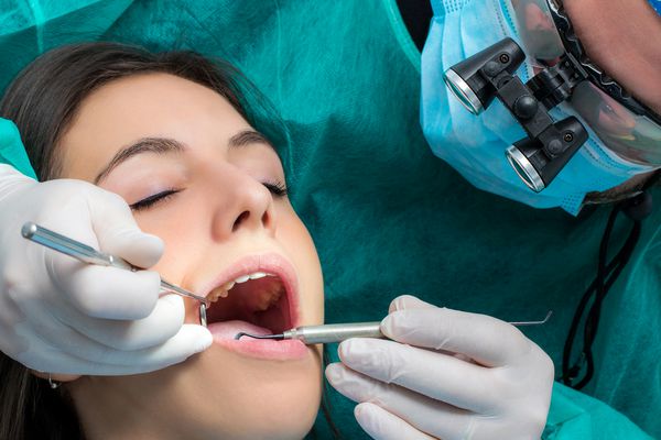 نزدیک دندانپزشک که دارای عینک ذره بین پزشکی است که روی بیمار زن کار می کند جراح در لباس سبز با آینه دهان و مقیاس دهنده