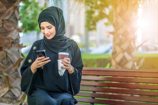 قهوه عرب و تلفن همراه بازرگانان عرب در حجاب در حال نگه داشتن قهوه در فضای باز و خواندن پیام از طریق تلفن همراه و نگاه کردن به دوربین هستند این زن در لباس ابایی سیاه پوشیده است