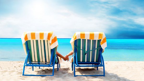نمای عقب یک زن و شوهر روی صندلی عرشه ای که در ساحل استراحت می کنند
