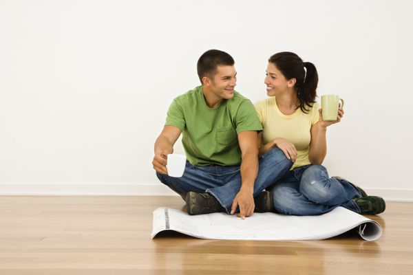 زوج جذابی که در کف خانه با فنجان های قهوه نشسته اند و برنامه های خانه را نگاه می کنند و به یکدیگر لبخند می زنند