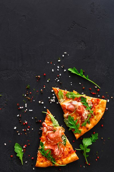 برش های پیتزا با prosciutto و سالاد موشک با ادویه جات ترشی جات در زمینه سیاه فضای کپی نمای بالا