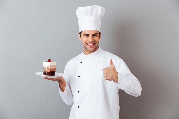 پرتره یك سرآشپز نر خنده دار كه در تخته نگهدارنده یونیفرم با تكه ای از كیك قرار گرفته و نشان دادن انگشت شست جدا شده از پس زمینه خاکستری