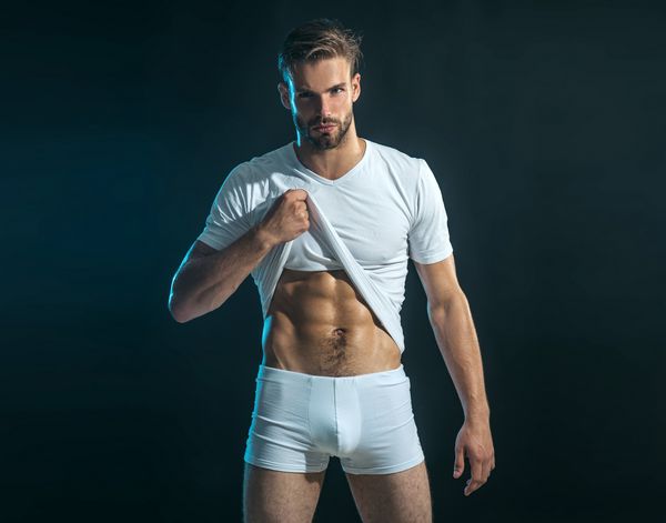 مرد در شلوار بوکس عضلات شکمی خود را نشان می دهد و تی شرت خود را با دست بالا می برد جوانی عضلانی در تی شرت سفید