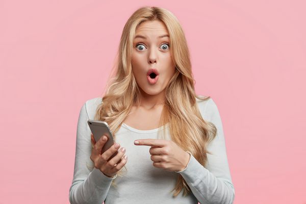 زن جوان با ظاهر دلپذیر با ظاهر وحشتناک در تلفن هوشمند به نظر می رسد اخبار تکان دهنده در صفحه وب جدا شده از پس زمینه صورتی را می خواند زن با تلفن دیجیتال نشان می دهد