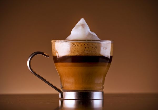 عکس نزدیک از نوشیدنی قهوه خوشمزه به نام caffe macchiato که اسپرسو با شیر و فوم است و شربت شکلات را برای اثر لایه ای به آن اضافه کرده است در یک لیوان شیشه ای سرو شده است