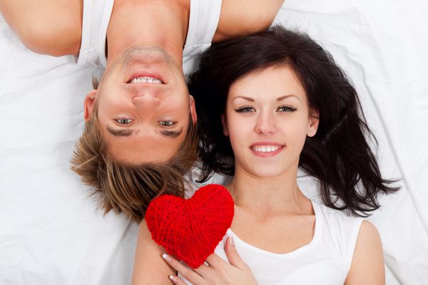 زن و شوهر دوست داشتنی جوان قلب قرمز را در کنار هم در یک تخت دراز می کشند لبخند به دنبال دوربین مفهوم عشق روز
