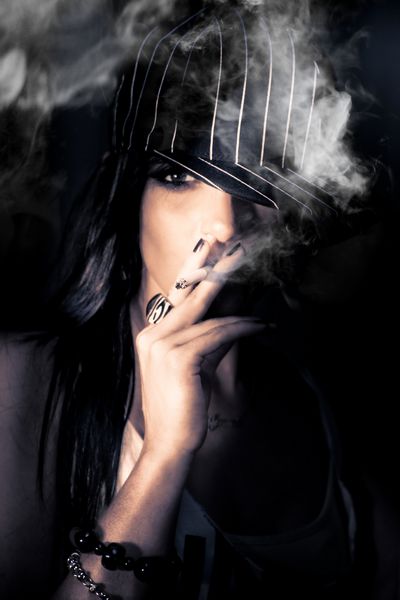 چهره یک زن شهری مرموز در کلاه راه راه ایستاده در سایه های ظلمت سیگار کشیدن سیگار در یک دود دودی مفهومی تمرکز بر روی خاکستر سیگار