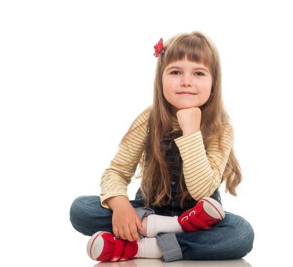 دختر کوچک ناز پوشیده از شلوار جین که روی زمین نشسته و روی زمینه سفید لبخند می زند