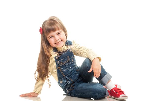 دختر کوچک ناز پوشیده از شلوار جین که روی زمین نشسته و لبخند می زند