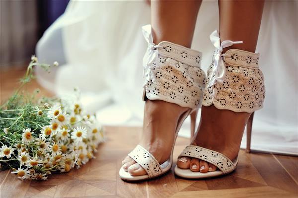 پاهای زنانه در صندل های سفید عروسی با دسته گل بابونه