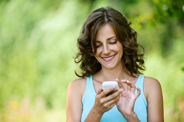 نمای نزدیک زن جوان جذاب با پیراهن آبی رنگ پیام را به تلفن همراه در مقابل رنگ سبز پارک تابستانی می خواند