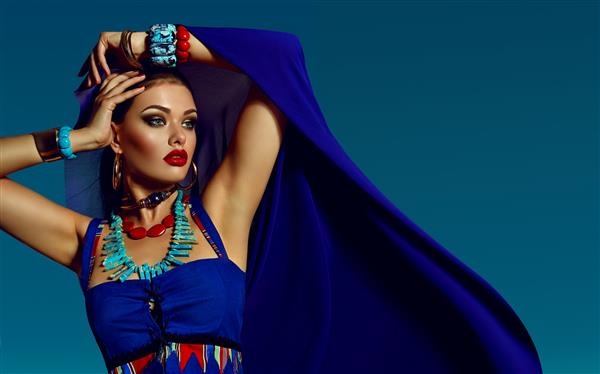 یک دختر جوان زیبا با لباس های آبی روشن و بسیاری از جواهرات در پس زمینه آسمان روشن دستبند گردنبند طلا مد زیبایی آرایش لوازم آرایشی سالن زیبایی سبک مراقبت شخصی