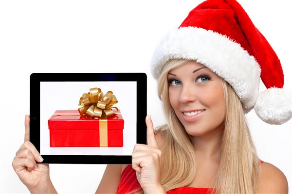 دختری بلوند با کلاه قرمز کریسمس در سال جدید یک ابزار لمسی پد لمسی رایانه لوحی را با جعبه هدیه بر روی صفحه در دست دارد