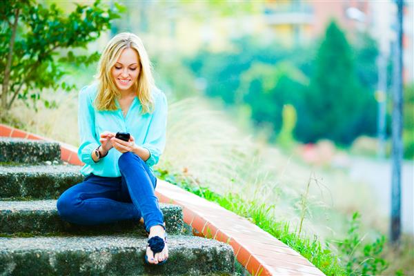 دختری خوشحال و خندان که روی پله ها نشسته و در تلفن همراه خود در پس زمینه پارک شهر پیامی می نویسد