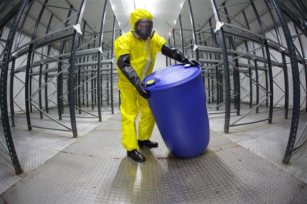 کارگر با لباس محافظ ماسک دستکش و چکمه در حال غلتیدن بشکه های مواد شیمیایی در انبار خالی - لنز چشم ماهی