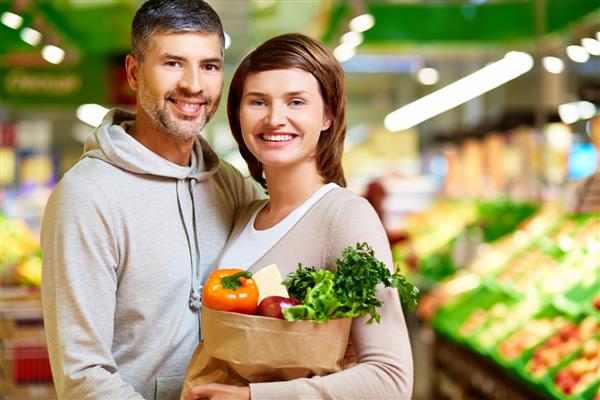 تصویری از یک زن و شوهر خوشحال با کیسه کاغذی پر از محصولاتی که در سوپر مارکت به دوربین نگاه می کنند