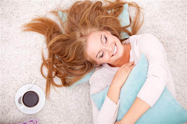 نمای بالایی از زن خندان دراز کشیده روی فرش با بالش و قهوه در خانه