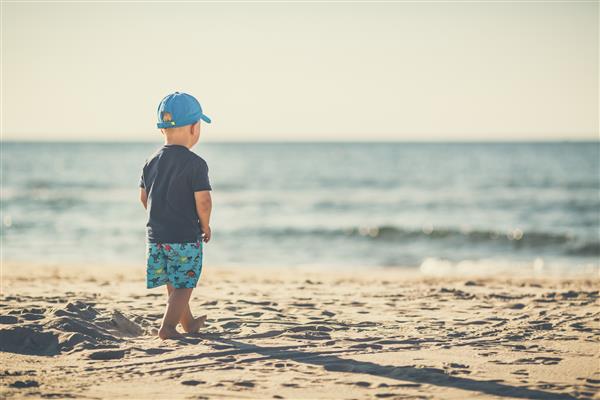 پسر نوپایی که در ساحل آفتابی قدم می زند کودک کوچک روی شن و ماسه راه می رود چشم انداز و چشم انداز زیبا از ساحل و اقیانوس الهام بخش
