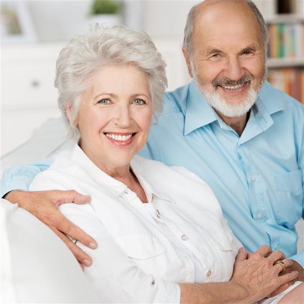 زن و شوهر سالخورده ای رمانتیک که در آغوش عاشقانه لبخند به دوربین می زنند و روی مبل اتاق نشیمن خود نزدیک یکدیگر نشسته اند