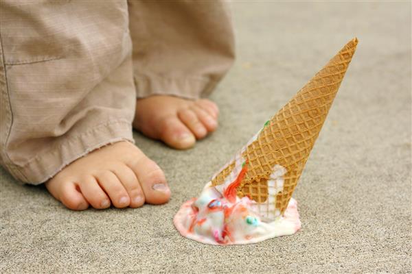 یک مخروط بستنی رنگی رنگین کمان افتاده به صورت وارونه در پیاده رو در پای کودک خردسال قرار دارد