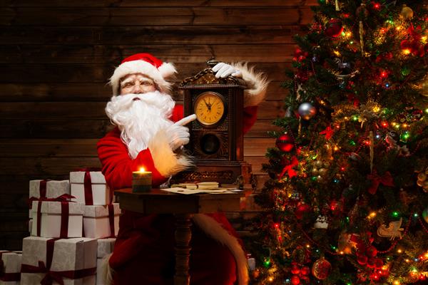 بابانوئل در فضای داخلی خانه چوبی و نشان دادن زمان در یک ساعت