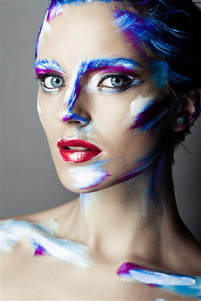 گریم هنری خلاقانه از یک دختر جوان با چشم های آبی ضربه های رنگی روی صورت و موهایش