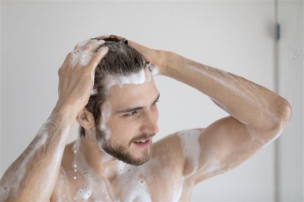 نزدیک مرد جوان برهنه قفقازی که موهای تمیز خود را با شامپوی طبیعی کف در حمام شستشو می دهد مرد هزاره دوش صبح می کند تمیز می کند در حمام بهداشت بدن مفهوم برنامه روزمره