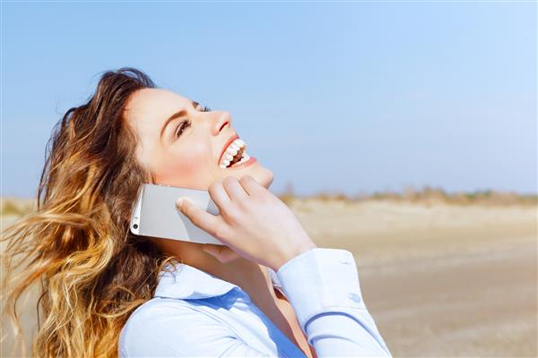 زن شاد با تلفن همراه کنار دریا می خندد