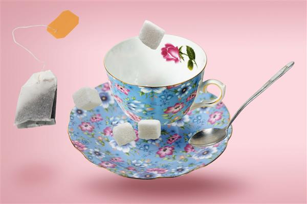 لیوان چای خالی تزئین شده آبی با نعلبکی کیسه چای قاشق و حبه های قند در حال پرواز روی سطح صورتی