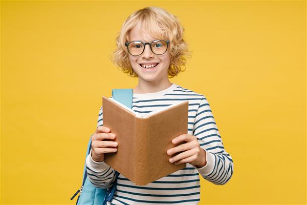 پسر بچه نوجوان پسر خنده دار و خنده دار 10 ساله در عینک کتانی راه راه و کوله پشتی دارای کتاب خواندن مدرسه است که در زمینه رنگ زرد تصویر کودک استودیو قرار دارد مفهوم آموزش
