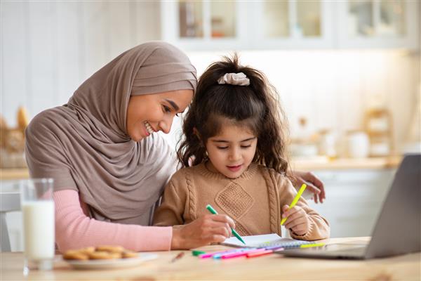 دوست داشتن مادر مسلمان با نقاشی با حجاب با دختر کوچولوی کودک پیش دبستانی اش در آشپزخانه خانواده اسلامی مبارک که پشت میز نشسته اند و با استفاده از مداد رنگارنگ در خانه با هم سرگرم می شوند فضای آزاد