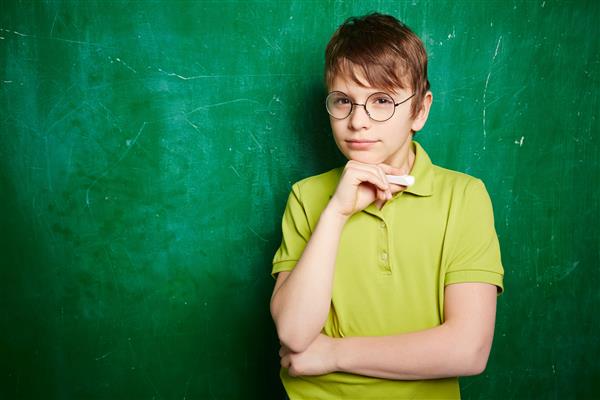 پرتره بچه مدرسه ای زیبا در عینک چشم که به دوربین کنار تخته سیاه نگاه می کند