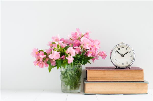 دکوراسیون منزل یکپارچه انبوهی از کتابها گلها و یک ساعت زنگ دار قدیمی در یک قفسه دیواری سفید