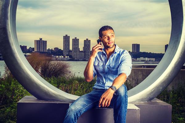 زندگی مدرن پیراهن آبی روشن شلوار جین آبی یک دانشجوی جوان خوش تیپ دانشگاه با ریش کوچک سبیل کنار رودخانه نشسته و با تلفن همراه صحبت می کند