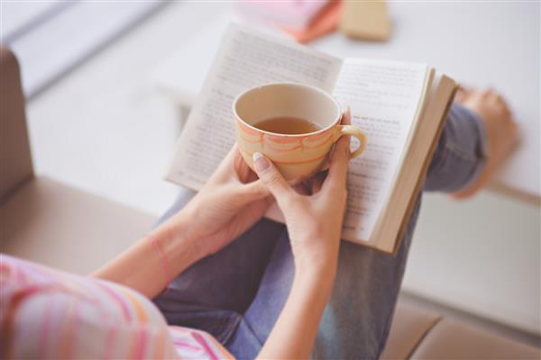 نمای نزدیک از دستان زن که لیوان چای را جلوی کتاب باز شده گرفته اند