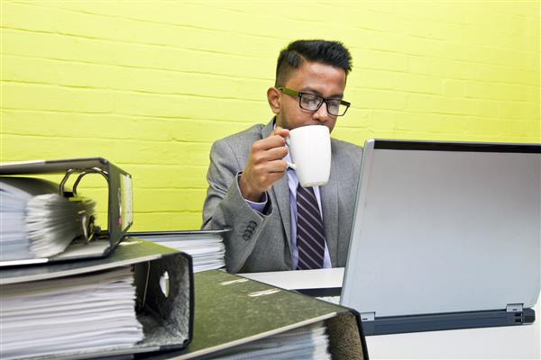 پرتره یک تاجر هندی که لیوان را در دست دارد و روی رایانه لپ تاپ خود پشت میز کارش کار می کند