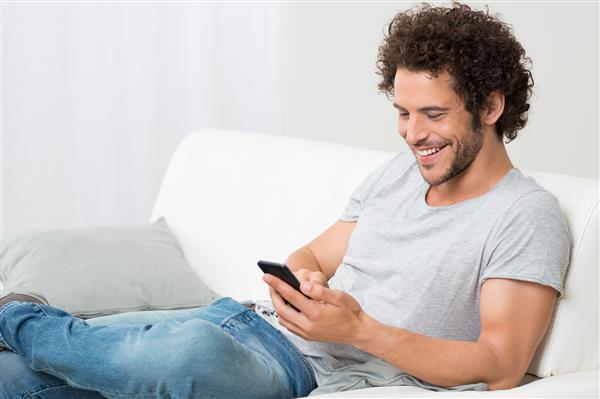مرد جوان مبارک که روی مبل استراحت می کند و به تلفن همراه نگاه می کند