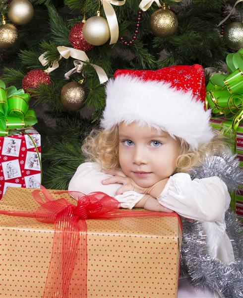 عکس زیبا از دختر کوچک ناز با موهای فرفری بور در کلاه سانتا و نشسته کنار درخت کریسمس تزئین شده با هدیه بزرگ