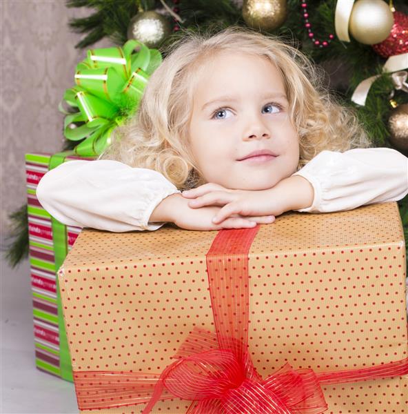 عکس زیبا از دختر بچه ناز با موهای فرفری بور که در کنار یک درخت کریسمس تزئین شده با هدیه بزرگ نشسته است