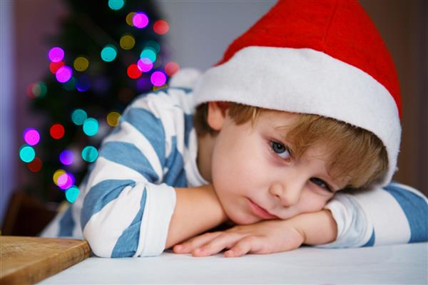 کودک کمی غمگین یا جدی با کلاه سانتا با درخت کریسمس و چراغ های زمینه