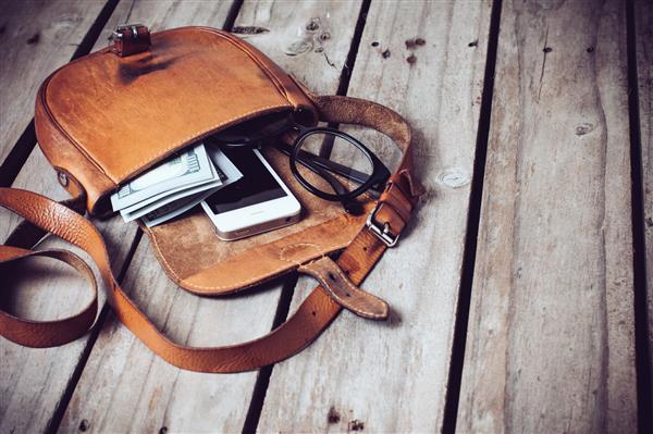 عینک نوری پول و تلفن هوشمند در کیف چرمی باز روی زمینه تخته چوبی