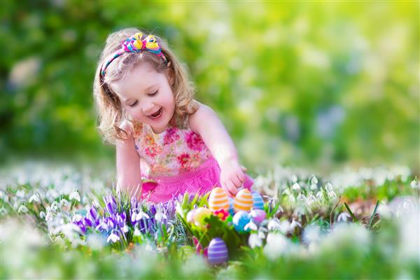 دختر نوپای فرفری با لباس تابستانی صورتی رنگ که با تخم مرغ های عید پاک